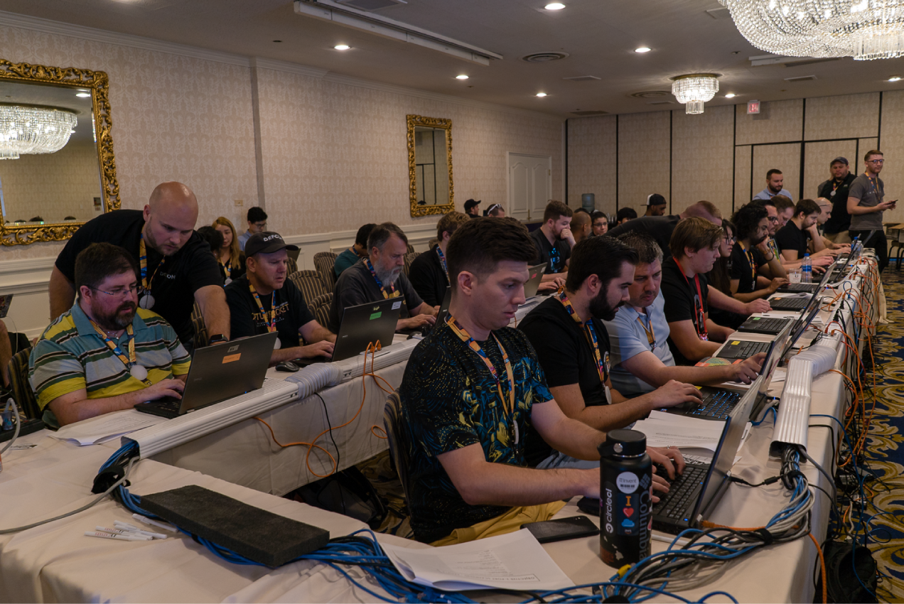 A room of hackers sitting at long tables with laptops.
