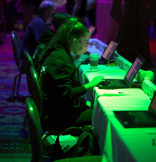 A female hacker wearing a mask sits at a table with others using a laptop at the Packet Hacking Village.