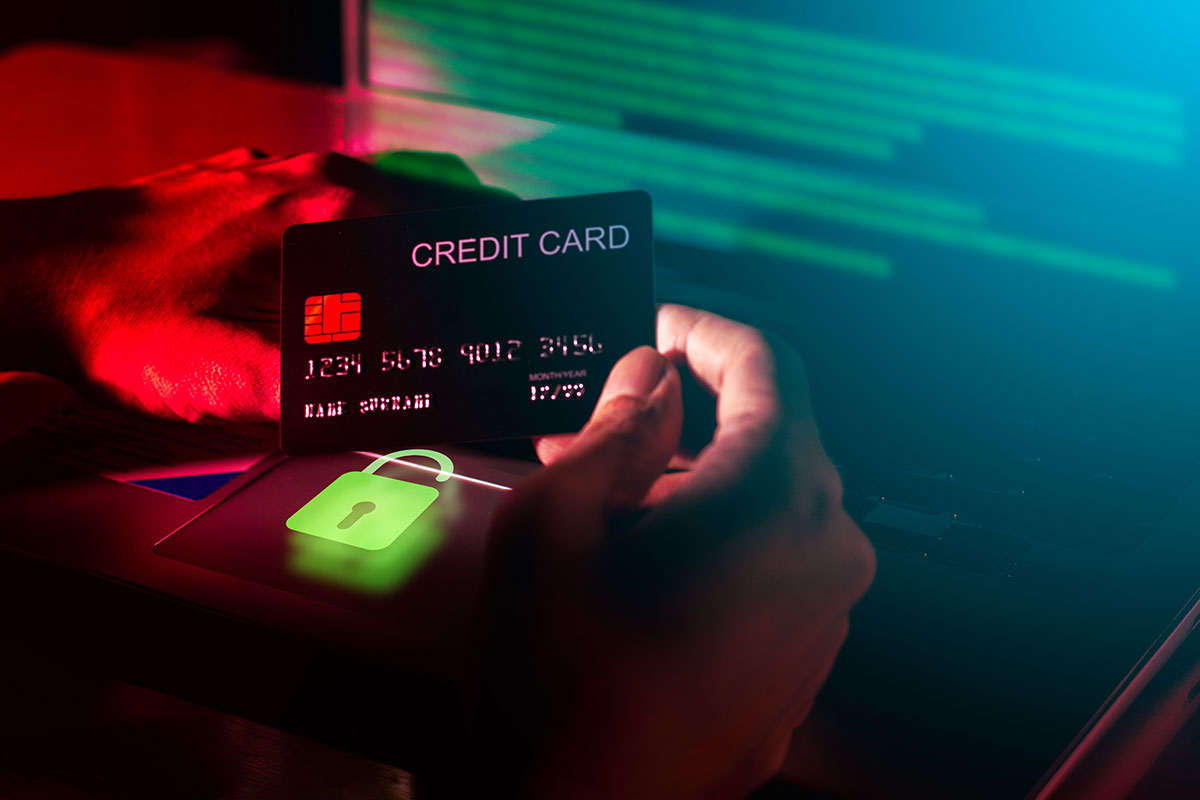 The hacker's hand is holding a credit card with a blurry computer background. 